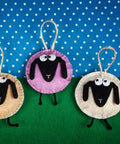 Three felt sheep ornaments with black faces on blue polka dot background, featuring colorful stitched edges and jute hangers.
