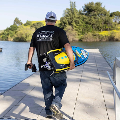 Person walking towards lake carrying Sonicwake RC Boat 48 Inch Self Righting Brushless 8S Ready To Run with controller