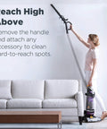 Side view of a person using a purple upright vacuum cleaner to reach high spots above a sofa.