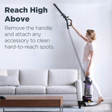 Side view of a person using a purple upright vacuum cleaner to reach high spots above a sofa.