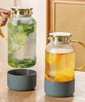 Two Piece Glass Pitcher Set Including Secure Glass Lid And Cushioned Anti Slip Silicone Base For Kitchen Counter Safe Serving Heavy Duty Water Jug Hassle Free Dispensing For Iced Coffee And Fresh Juice on wooden table, filled with lime and orange slices.