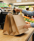 Person holding Big 14 By 10 By 16 Inches Brown Paper Bags With Handles for takeout and shopping at a grocery checkout.