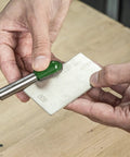 Hands using All In One Credit Card Diamond Sharpening Kit With 180 300 Grit And 600 1000 Grit Stones Lapping Fluid Non Slip Mat Cleaning Block And Sharpening Guide To Get Your Blades Sharp Like A Boss showing 600 grit stone and tool on wooden table.