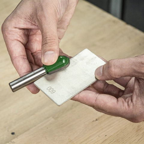 Hands using All In One Credit Card Diamond Sharpening Kit With 180 300 Grit And 600 1000 Grit Stones Lapping Fluid Non Slip Mat Cleaning Block And Sharpening Guide To Get Your Blades Sharp Like A Boss showing 600 grit stone and tool on wooden table.