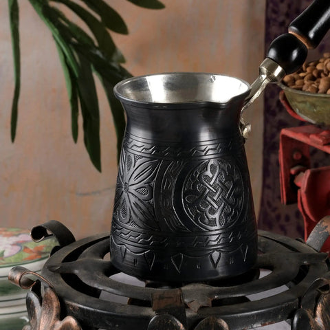 Black engraved Turkish coffee pot with metallic interior and wooden handle on a decorative stovetop, rich cultural patterns visible.