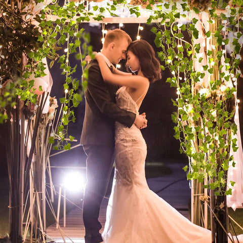 Couple embracing under arch of 33 Ft Ivy Garland With 100 LED Fairy Lights For Home Decor Living Room And Bedroom Wall Art Aesthetic Office Lighting Wedding Party Farmhouse Dorm Outdoor Garden Glow, creating a romantic ambiance.