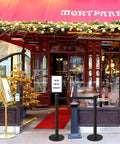 Image of a stylish restaurant entrance with Retractable Belt Stanchions 3 Pack for Crowd Control Barriers With Ropes and Poles Black Six Point Six Feet Tall Durable Stainless Steel Posts With Sign Frame Holder used for effective queue management, featuring a decorative awning and festive decorations.