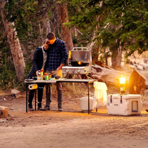 Couple using a folding tailgate table with grill rack for outdoor camping and grilling. Features 5-foot portable prep station.