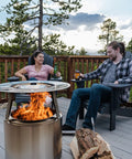 Ranger Heat Deflector Boosts Outdoor Fire Pit Warmth With Angled Design visible in use on a deck with two people enjoying the fire