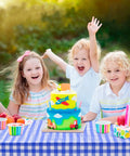 Children celebrating at a picnic with a vibrant birthday cake on a blue checkered tablecloth outdoors.