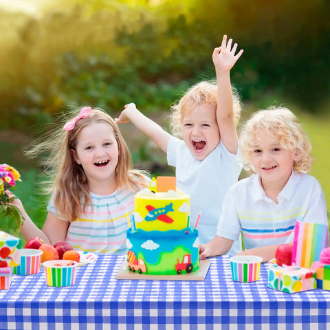 Children celebrating at a picnic with a vibrant birthday cake on a blue checkered tablecloth outdoors.