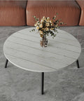 Round table with white gray wood-like surface matching wallpaper theme, placed in a living room with couch and vase of flowers.