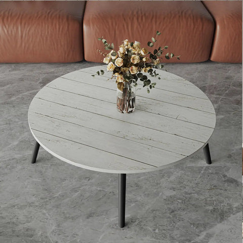 Round table with white gray wood-like surface matching wallpaper theme, placed in a living room with couch and vase of flowers.