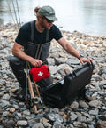Red NANUK 903 waterproof first aid box, held by a fisherman beside an open black case on a rocky riverbank.