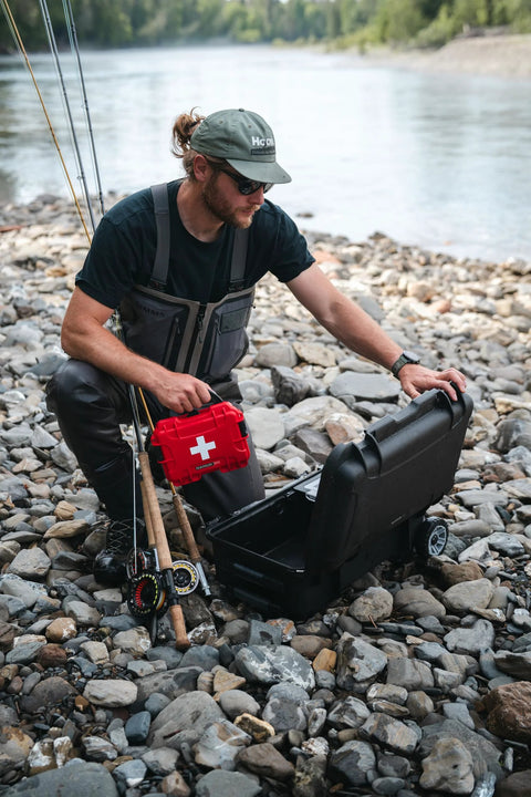Red NANUK 903 waterproof first aid box, held by a fisherman beside an open black case on a rocky riverbank.
