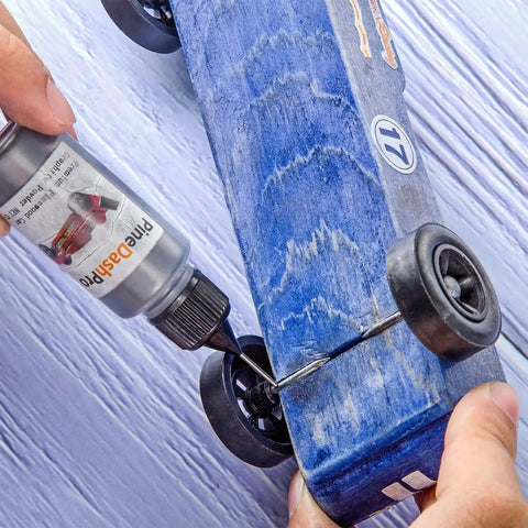 Applying graphite lubricant from the Two Size Pinewood Derby Weights Kit With Twelve Piece Weight Set Including Four Half Ounce And Eight Quarter Ounce Weights And Half Ounce Graphite Lubricant Powder For Derby Wheels And Axles To Boost Car Speed to a blue derby car wheel