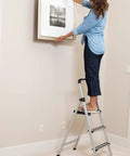 Woman hanging wall art while standing on Lightweight Aluminum 3 Step Ladder With Wide Non Slip Steps Foldable Stepladder For Kitchen Pantry Closets And Home Office Space Saving Indoor Use Handy Portable Step Stool Durable Silver Finish