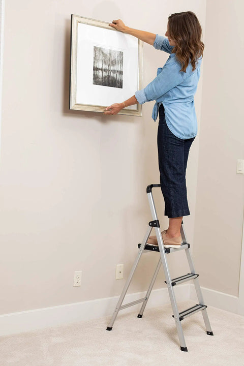 Woman hanging wall art while standing on Lightweight Aluminum 3 Step Ladder With Wide Non Slip Steps Foldable Stepladder For Kitchen Pantry Closets And Home Office Space Saving Indoor Use Handy Portable Step Stool Durable Silver Finish