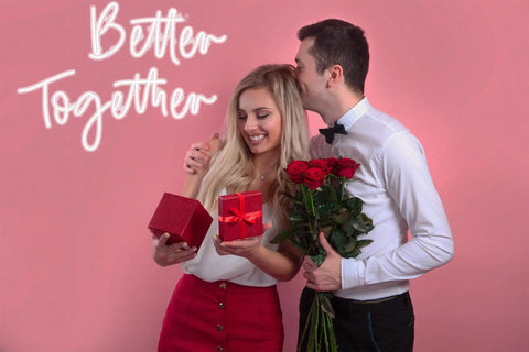 Couple giving gifts with "Better Together" neon sign on pink background