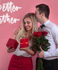 Couple with roses and gifts in front of 'Better Together' neon sign on pink wall