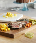 Gourmet platter with sliced seared tuna, diced mango, avocado, and carrot julienne on a wooden board, cake in background.