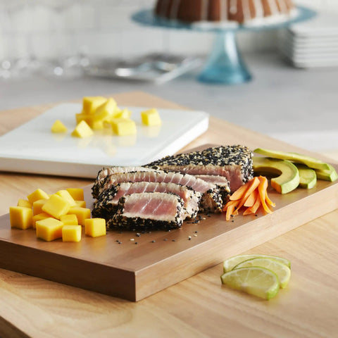Gourmet platter with sliced seared tuna, diced mango, avocado, and carrot julienne on a wooden board, cake in background.