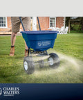 Estate Style Broadcast Spreader With 65 Lb Capacity And Powder Coated Steel Frame spreading seeds on lawn with pneumatic tires.