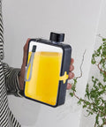 Person holding Flat Square Water Bottle For On The Go Hydration, Leak Proof And Drop Proof, black exterior with yellow liquid inside.
