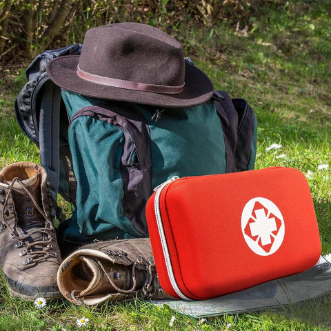 Front view of red emergency first aid kit with white star-of-life symbol beside green backpack and hiking boots.
