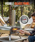 A man using a 16 Inch Multi-Fuel Outdoor Pizza Oven For Wood Fired Or Gas Cooking Reaches 1112 Fahrenheit, Stainless Steel Portable Pizza Cooker For Parties, 2-In-1 System With Burners Sold Separately at a campsite, featuring a visible glass panel and built-in thermometer.