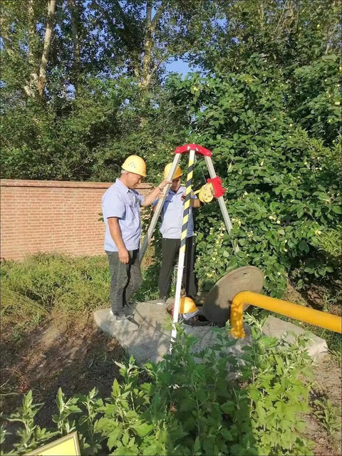 Two workers using Rugged Confined Space Tripod Rescue Rig Featuring Heavy Duty Winch Choices next to a manhole outdoors.