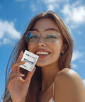 Front view of a smiling woman outdoors holding a Lulumonster anti-fog lens wipe packet for glasses (150 pre-moistened).