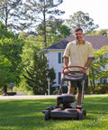Person mowing a lawn with a push lawn mower in a residential neighborhood, surrounded by trees and a white house in the background.