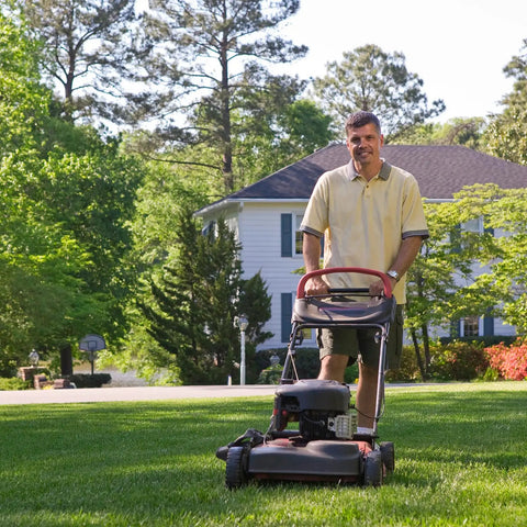 Person mowing a lawn with a push lawn mower in a residential neighborhood, surrounded by trees and a white house in the background.