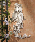 Brianna The Summer Breeze Fairy Wall Sculpture Hand Cast Stone Resin Garden Decor With Hand Painted Faux Stone Finish Whimsical Indoor Outdoor Wall Art Featuring Elegant Wings And Flowing Tresses mounted on a peach wall beside green vines and purple flowers.