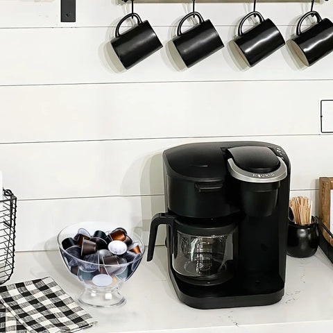Clear Coffee Pod Holder For K Cups And Vertuo Capsules next to a coffee maker on a kitchen countertop with black mugs above.