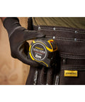 Gloved hand holding Fatmax Autolock Tape Measure 8m Metric Only Yellow Black, Reverse Lock For One-Handed Control, in tool belt.