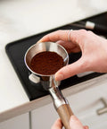 Badass Non Slip Silicone Tamping Mat For Dual Boiler Espresso Machines includes a hand holding a portafilter with ground coffee.