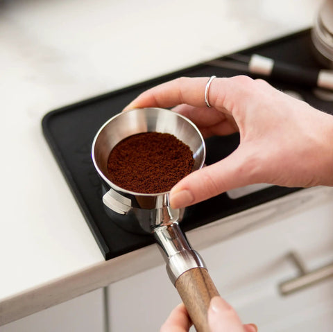 Badass Non Slip Silicone Tamping Mat For Dual Boiler Espresso Machines includes a hand holding a portafilter with ground coffee.
