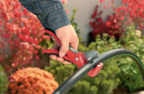Drip Tubing Cutter And Punch Gun Kit For 1 Inch Flexible Tubing cutting irrigation pipe in garden setting with red-handled tool.