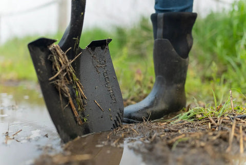 Short-Handle Digging Shovel With Heavy Duty Carbon Steel Build in use, showing serrated edge in muddy ground, rubber boot visible