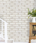 White and beige faux brick wall with greenery vase beside staircase, showcasing Peel And Stick Faux Brick Wallpaper Self Adhesive White And Beige Roll 17.7 By 472.4 Inches Removable Wall Covering For Bedroom And Laundry Room Accent Walls Easy Install No Glue Needed.