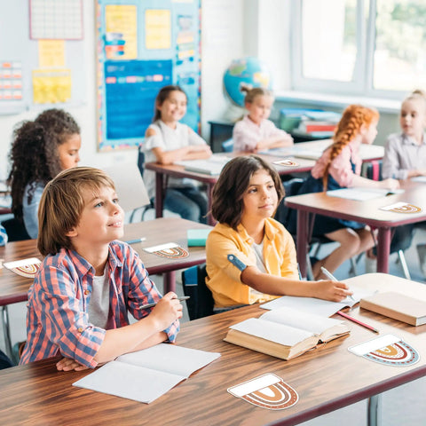 Students in a classroom with Rainbow Cutouts For Classroom Decor, 45 Pcs Boho Rainbow Name Tags With 100 Glue Dots on desks