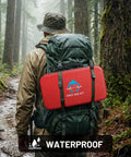 Back view of a red waterproof first aid kit attached to a green hiking backpack with straps in a forest