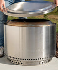 Person placing Yukon Lid Fire Pit Cover Premium 304 Stainless Steel Round Tabletop Accessory on a fire pit