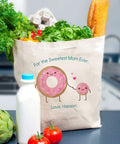 Canvas tote bag with 'For the Sweetest Mom Ever' and cartoon donuts, filled with groceries including lettuce, bread, and milk.