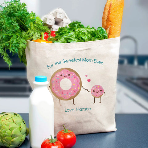 Canvas tote bag with 'For the Sweetest Mom Ever' and cartoon donuts, filled with groceries including lettuce, bread, and milk.