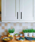 Kitchen setting with 30 Pack Solid 5 Inch Square Cabinet Handles Matte Black Pulls for kitchen cabinets, shown installed on white cabinets above countertop with plants.