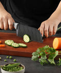 Front view of crinkle-cut knife with ribbed dark blade and gray rubber handle slicing cucumber on a wooden board