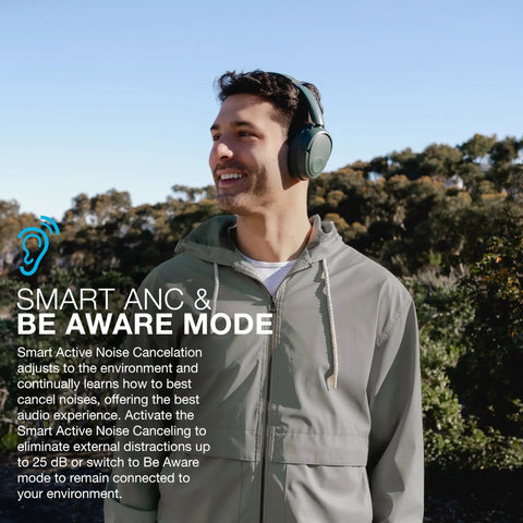 Man wearing JBuds Lux ANC Over Ear Headphones With Wireless Active Noise Cancellation outdoors, smiling under clear blue sky.